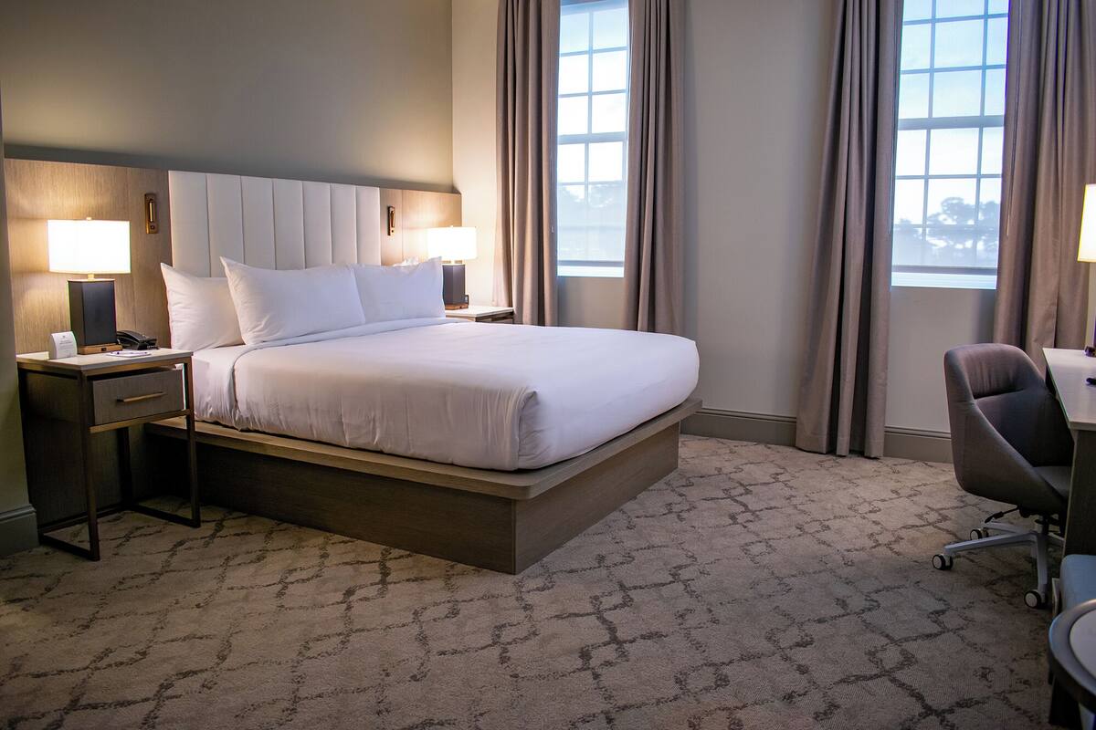 A hotel room with a large bed, two bedside tables with lamps, a desk with a chair, and a view of the outdoors through the windows.