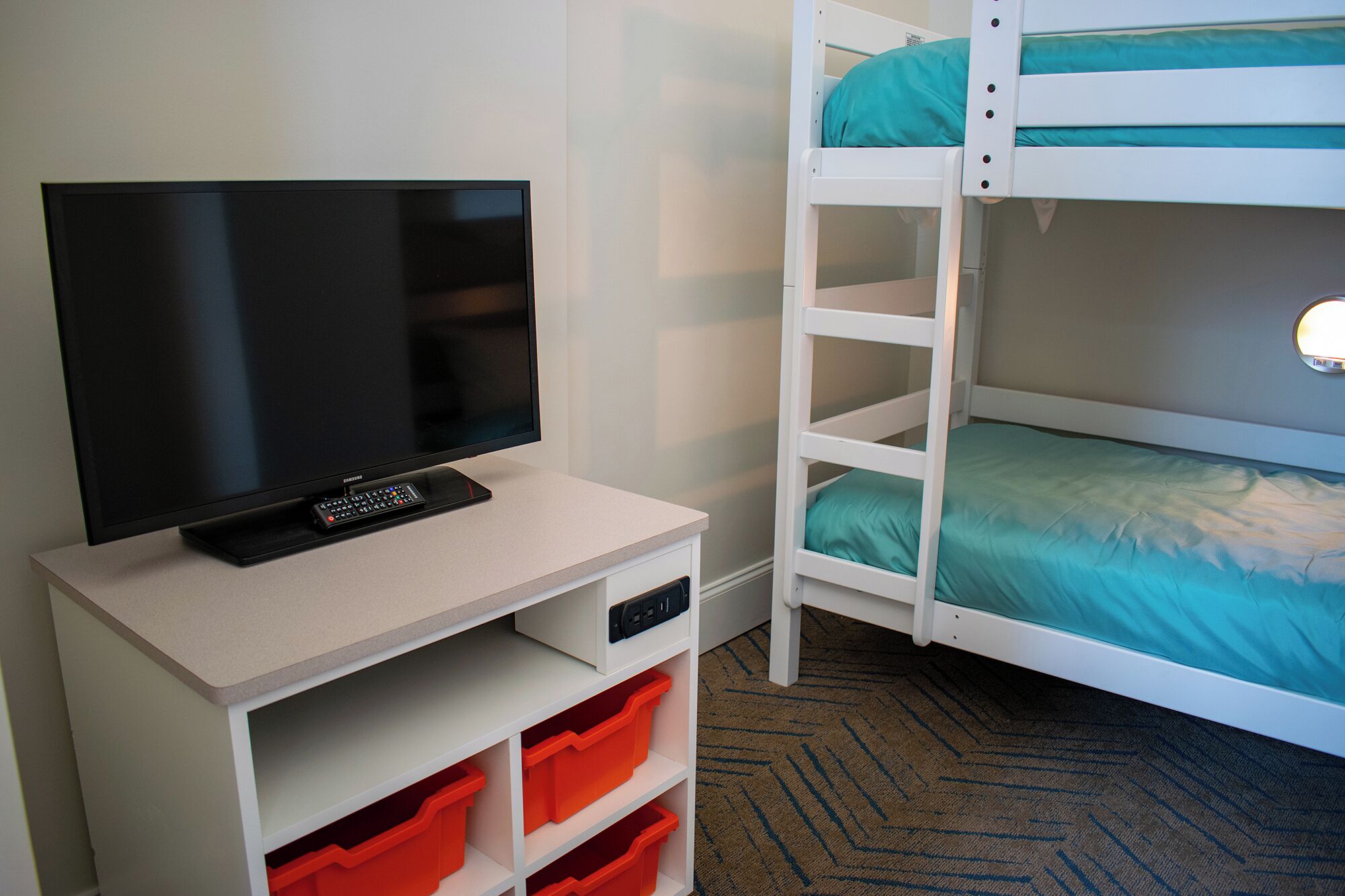 Una habitación con televisión sobre un mueble blanco, una litera con ropa de cama color turquesa y contenedores de almacenamiento naranja.