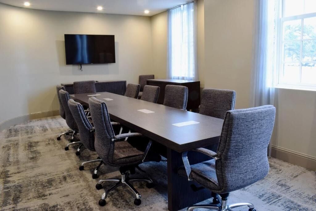A conference room with a long table, six chairs, a flat-screen TV, and a large window.