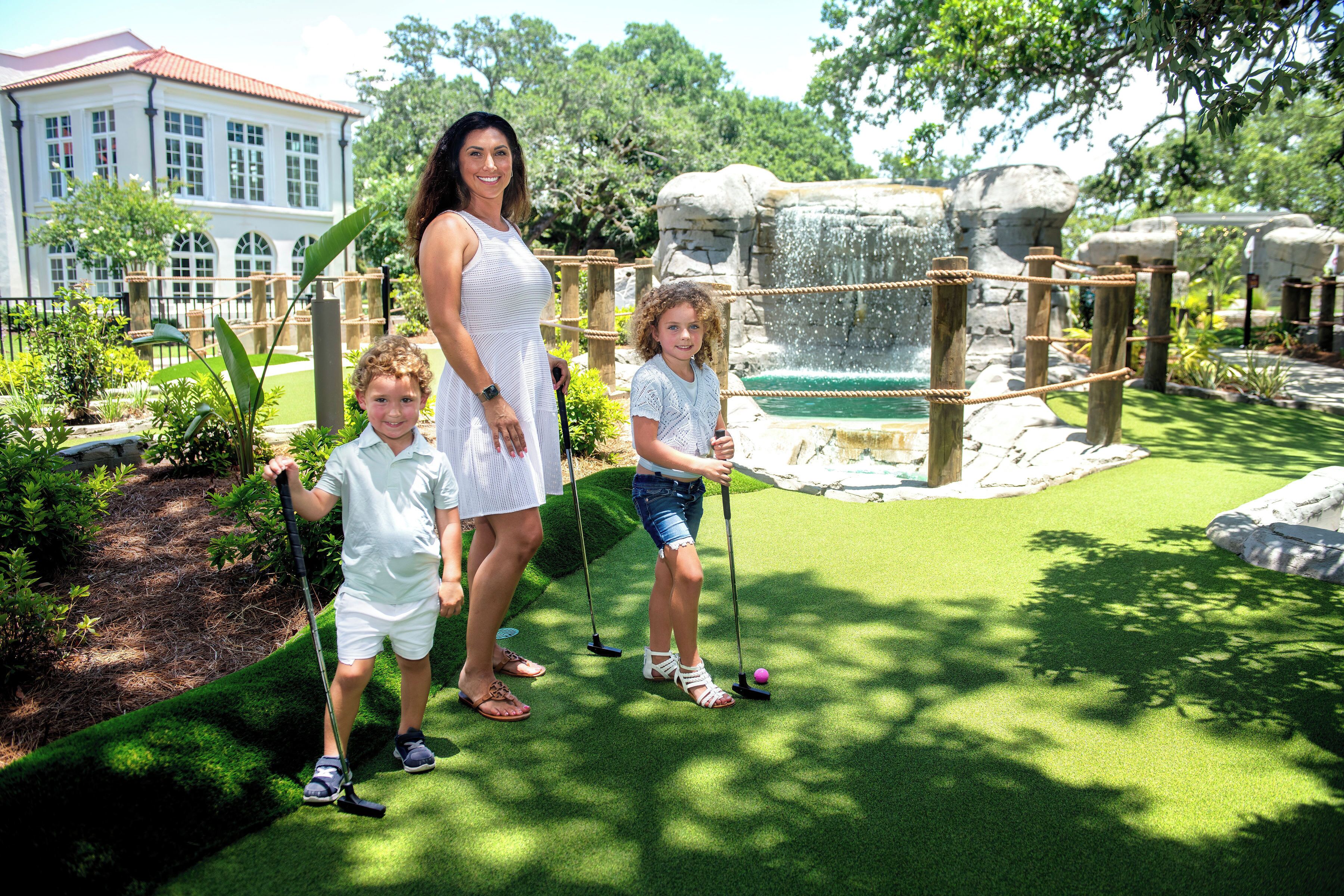 A woman and two children playing croquet in a park with a water feature and a building in the background.