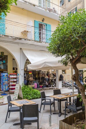 Um café ao ar livre com mesas e cadeiras, uma placa e uma loja com bolsas e acessórios.