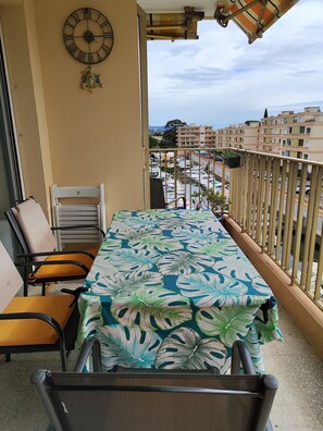 A balcony with a table covered in a tropical leaf tablecloth, chairs, a clock, and a view of a marina and buildings.