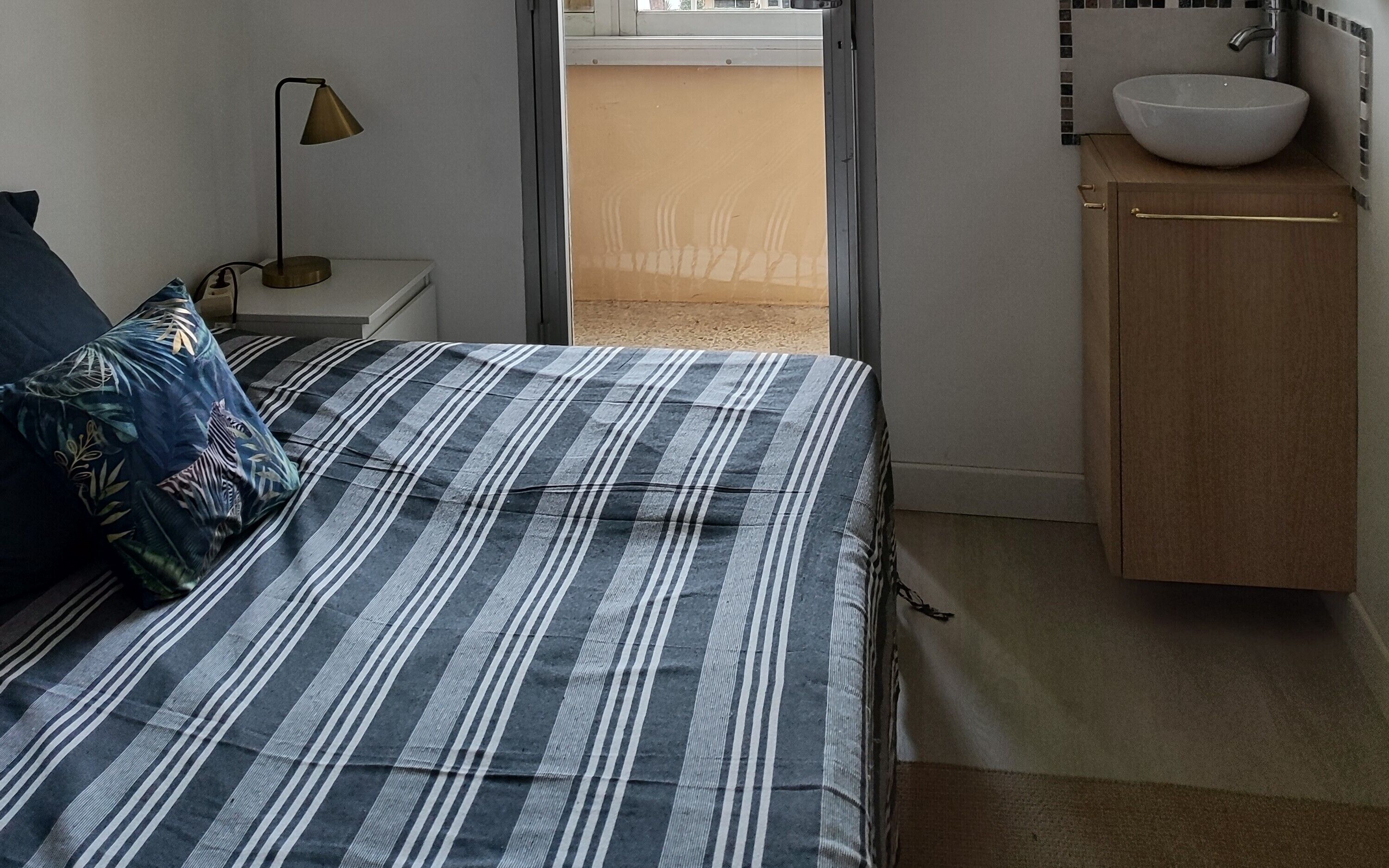 A bedroom with a striped bedspread, a bedside lamp, a wooden cabinet with a sink, and a view through a window.