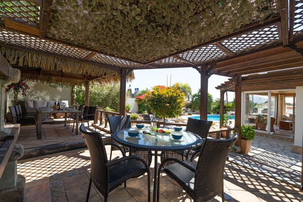 Un espace extérieur abrité, aménagé pour les repas, avec des meubles en osier, une table en verre et une vue sur une piscine et la verdure.