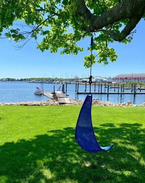 A clear view of a marina with boats docked, a grassy area with a hanging chair, and a building with 'MYSTIC SHIPYARD' signage in the background. - Harbor View Landing (Mystic)