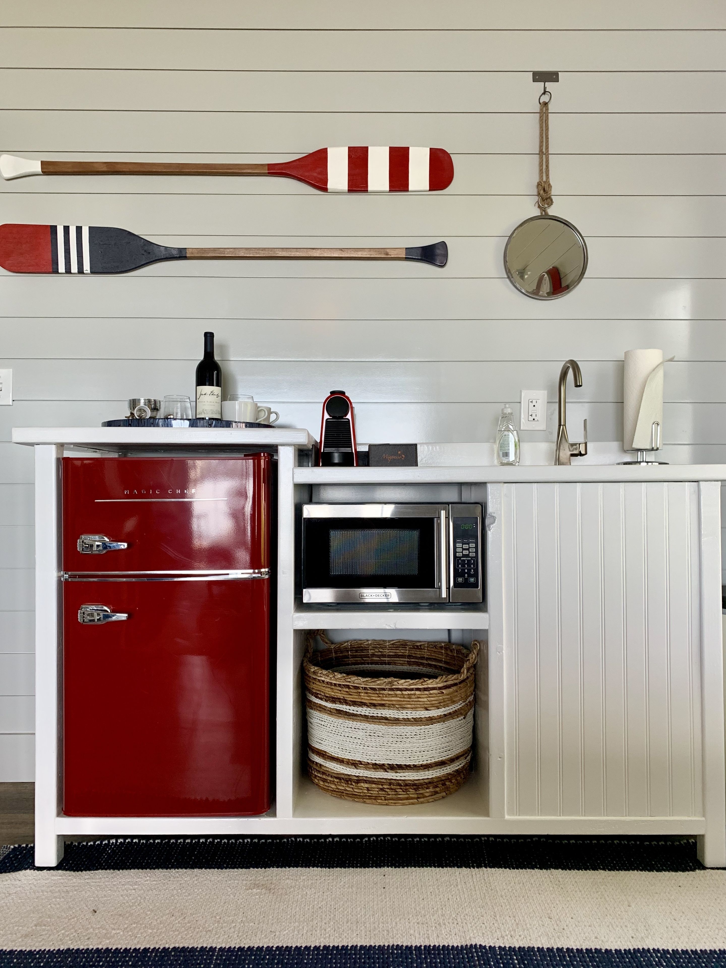 Une cuisine avec un réfrigérateur rouge, un four à micro-ondes et un évier blanc. On y trouve des pagaies en bois et un miroir rond accroché au mur.