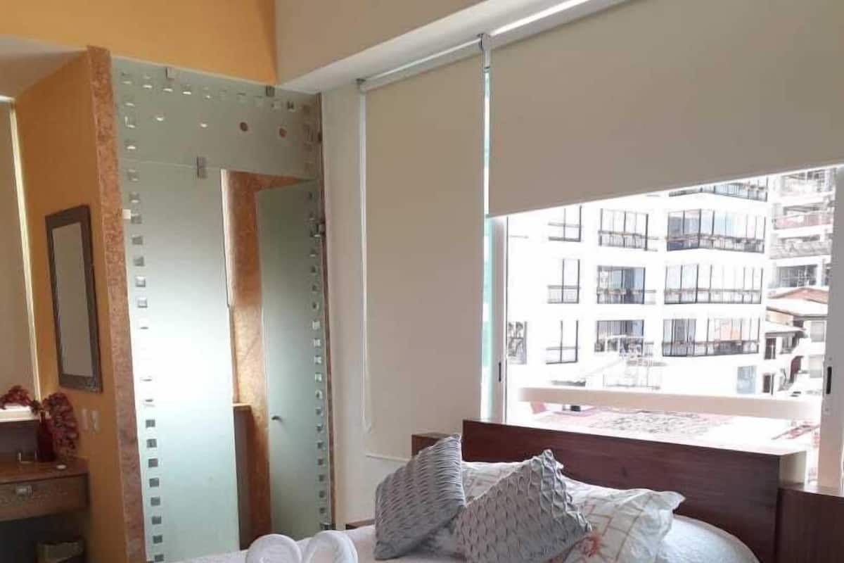 Hotellihuone, jossa on sänky, yöpöytä, tuoli ja ikkuna, josta on näkymä rakennuksiin.