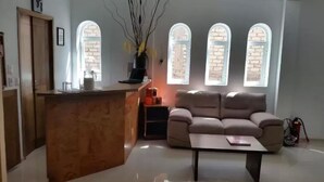 Una sala de estar con sofá, una mesa de centro, un mostrador de recepción de madera y un jarrón con ramas. Hay ventanas con diseños de ladrillo y un extintor en el suelo.