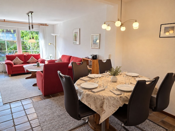 Un comedor con una mesa redonda dispuesta para cuatro personas, rodeada de sillas negras. En el fondo, un sofá rojo, y una ventana con cortinas.