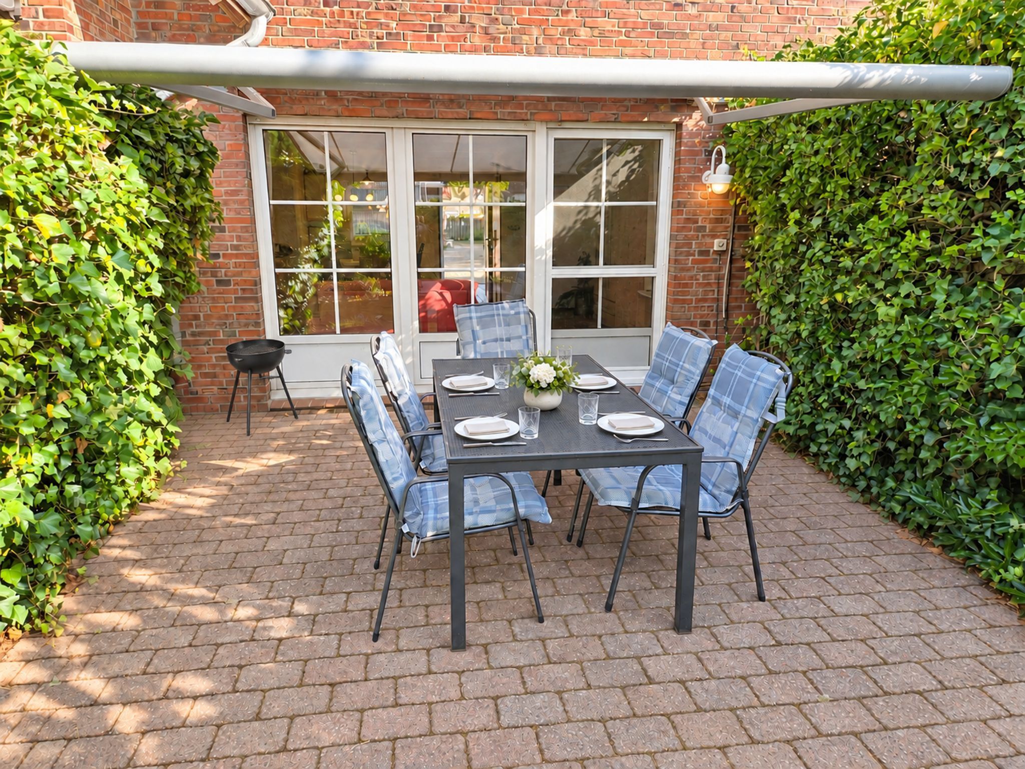 Une terrasse avec une table dressée pour quatre personnes, entourée de chaises, d’un barbecue et d’une fenêtre donnant sur un jardin.