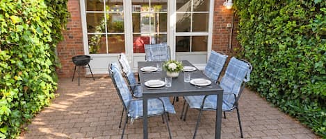 Eine Terrasse mit Tisch für vier Personen, umgeben von Stühlen, einem Grill und einem Fenster mit Blick auf einen Garten.
