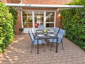 Une terrasse avec une table dressée pour quatre personnes, entourée de chaises, d’un barbecue et d’une fenêtre donnant sur un jardin.