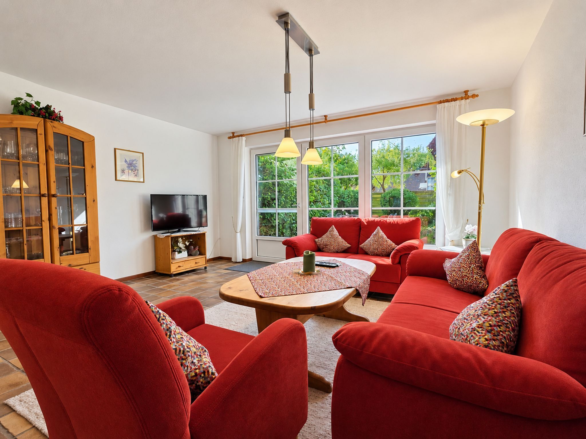 Een woonkamer met een rode zithoek, een houten kast, een televisie en een groot raam met uitzicht op groen.