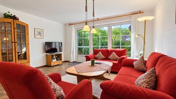 A living room with a red sofa set, a wooden cabinet, a television, and a large window with a view of greenery.