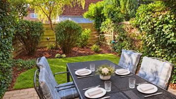 An outdoor patio with a table set for two, surrounded by greenery and a wooden fence.