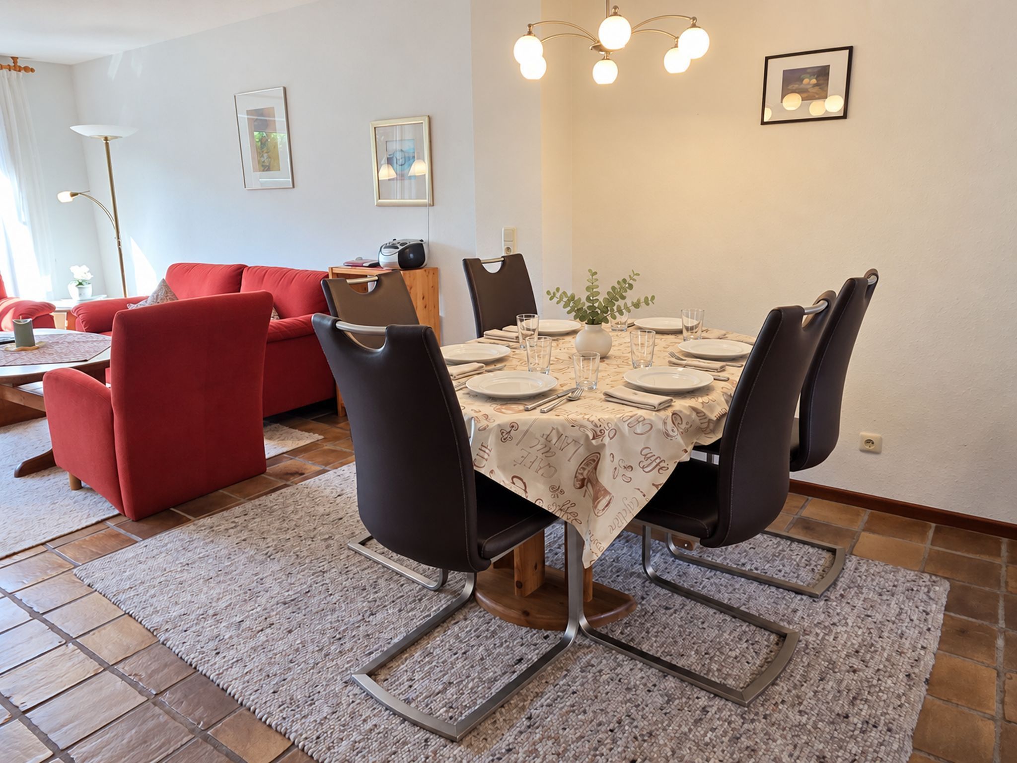 A dining area with a table set for four, surrounded by chairs, a red sofa, and a lamp in the background.