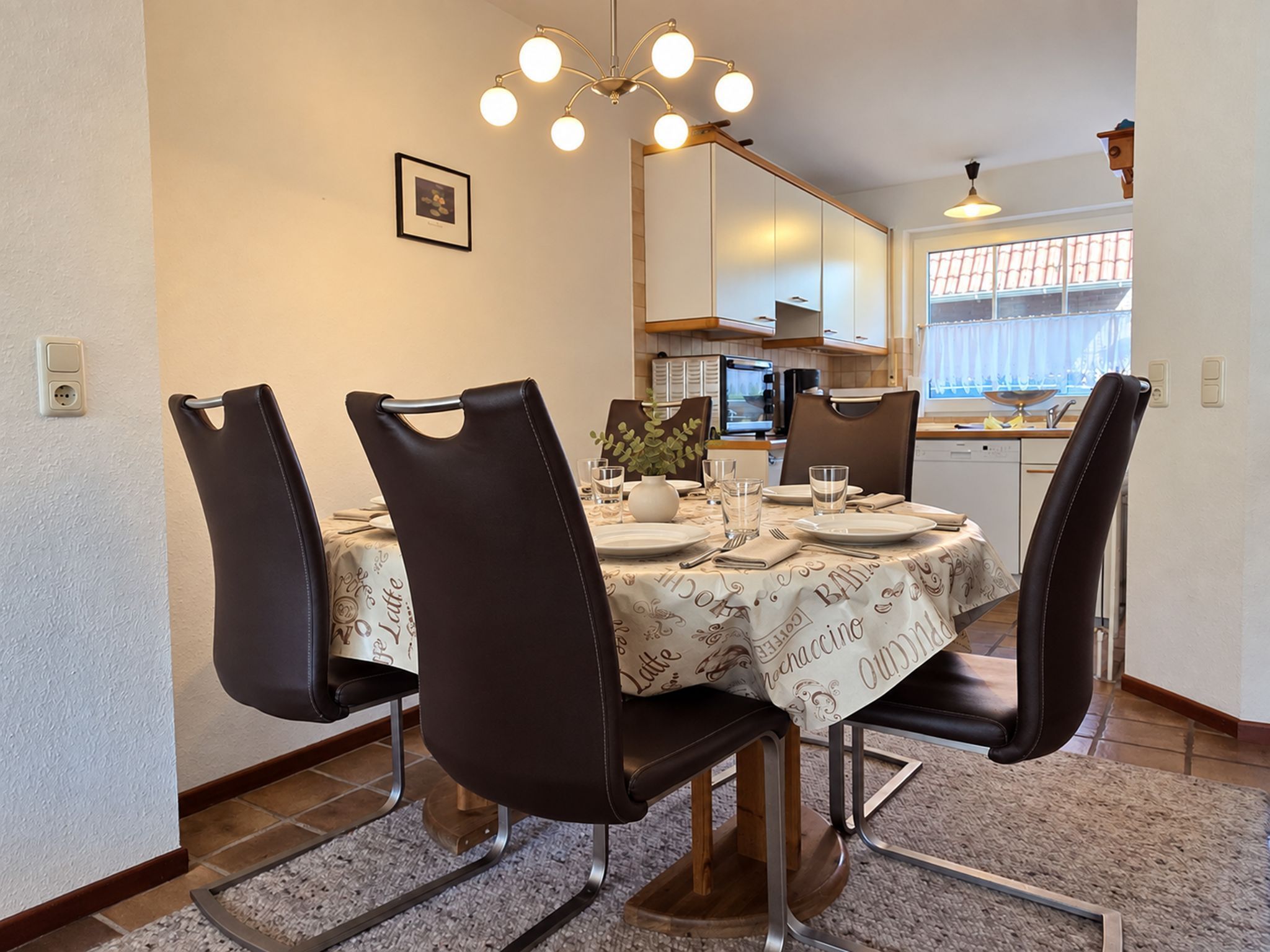A dining area with a round table, four chairs, a chandelier, and a kitchen in the background.