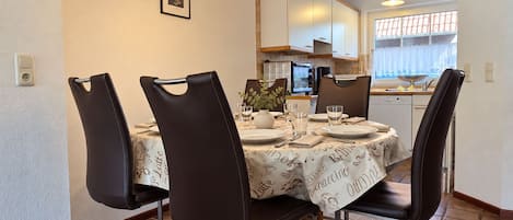 A dining area with a round table, four chairs, a chandelier, and a kitchen in the background.