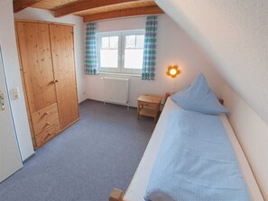 A bedroom with a wooden wardrobe, a small bedside table, a bed with a blue pillow, a window with curtains, and a wall-mounted light fixture.