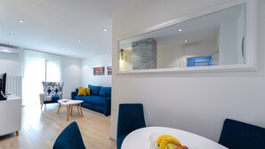 Una sala de estar moderna con un sofá azul, una mesa de comedor blanca y un bol de frutas, y un espejo grande sobre la mesa.