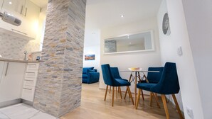 Cocina moderna con armarios blancos, pared decorativa de piedra y zona de comedor con sillas azules y una mesa pequeña.