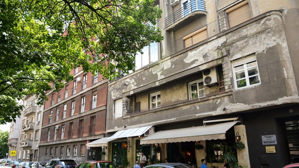 Vista de la calle con coches estacionados, un edificio con balcón y un restaurante con terraza.