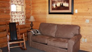 A log cabin interior with a brown leather sofa, a rocking chair, a framed picture on the wall, and a window with floral curtains. - Rand Creek Ranch (Cody)