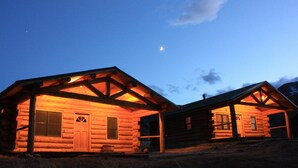 A log cabin with a porch and a small attached structure, both illuminated by warm lighting. - Rand Creek Ranch (Cody)