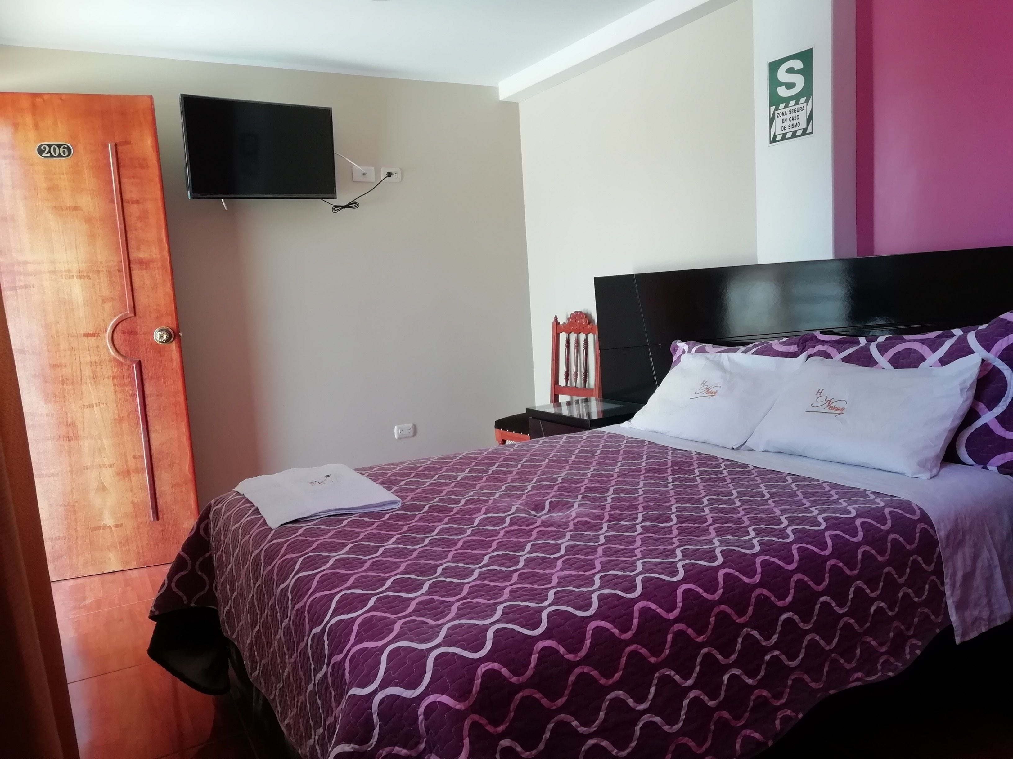 A hotel room with a bed, a television mounted on the wall, a wooden door, and a sign indicating a secure zone.