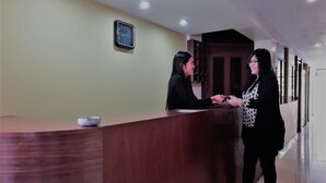 Zona de recepción con un mostrador de madera, un reloj en la pared y un pasillo con puertas y ventanas.