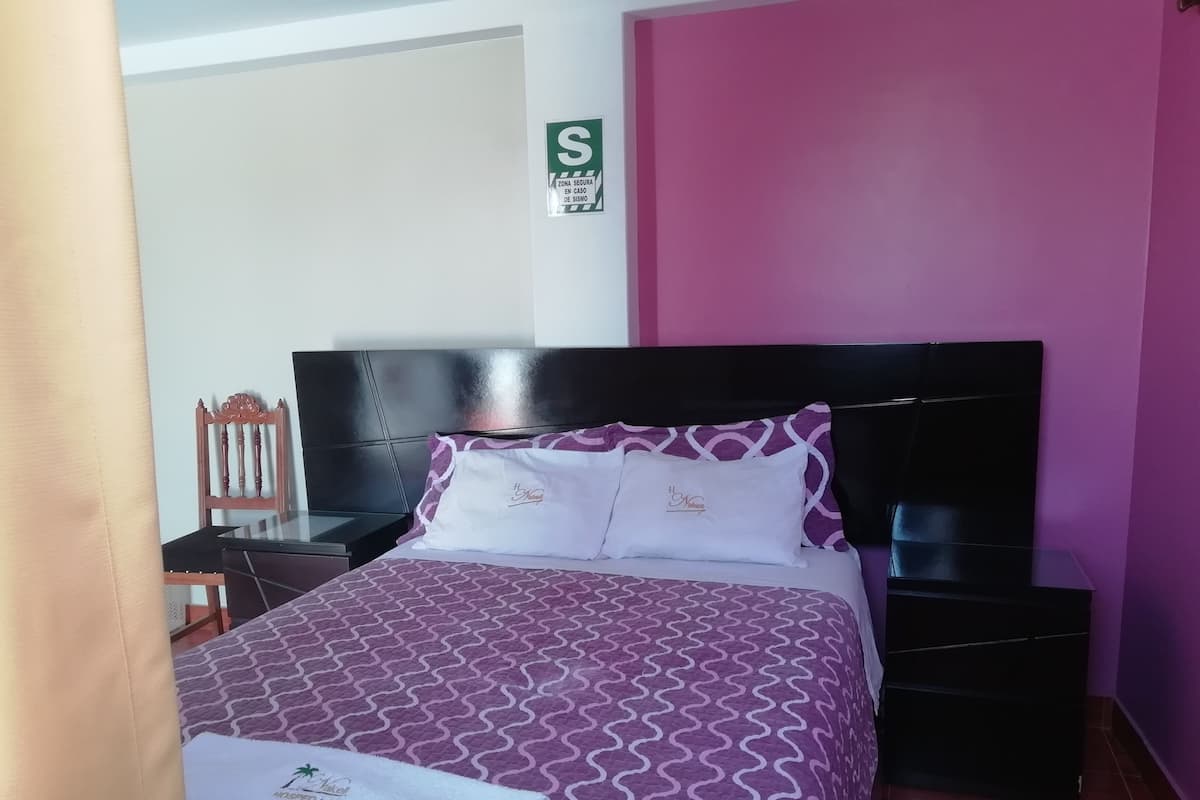 Cama bem arrumada com colcha e almofadas estampadas, uma cadeira de madeira, um criado-mudo e uma placa indicando zona segura.