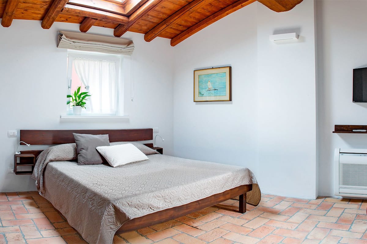 Un dormitorio con cama de madera, mesita de noche, ventana con planta, un cuadro en la pared y televisión en la pared.