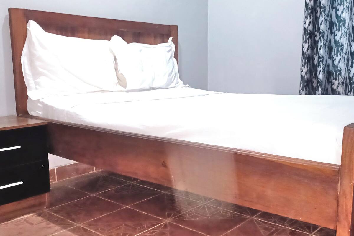 Una cama de madera con ropa de cama blanca, una mesita de noche con cajones y una cortina estampada.