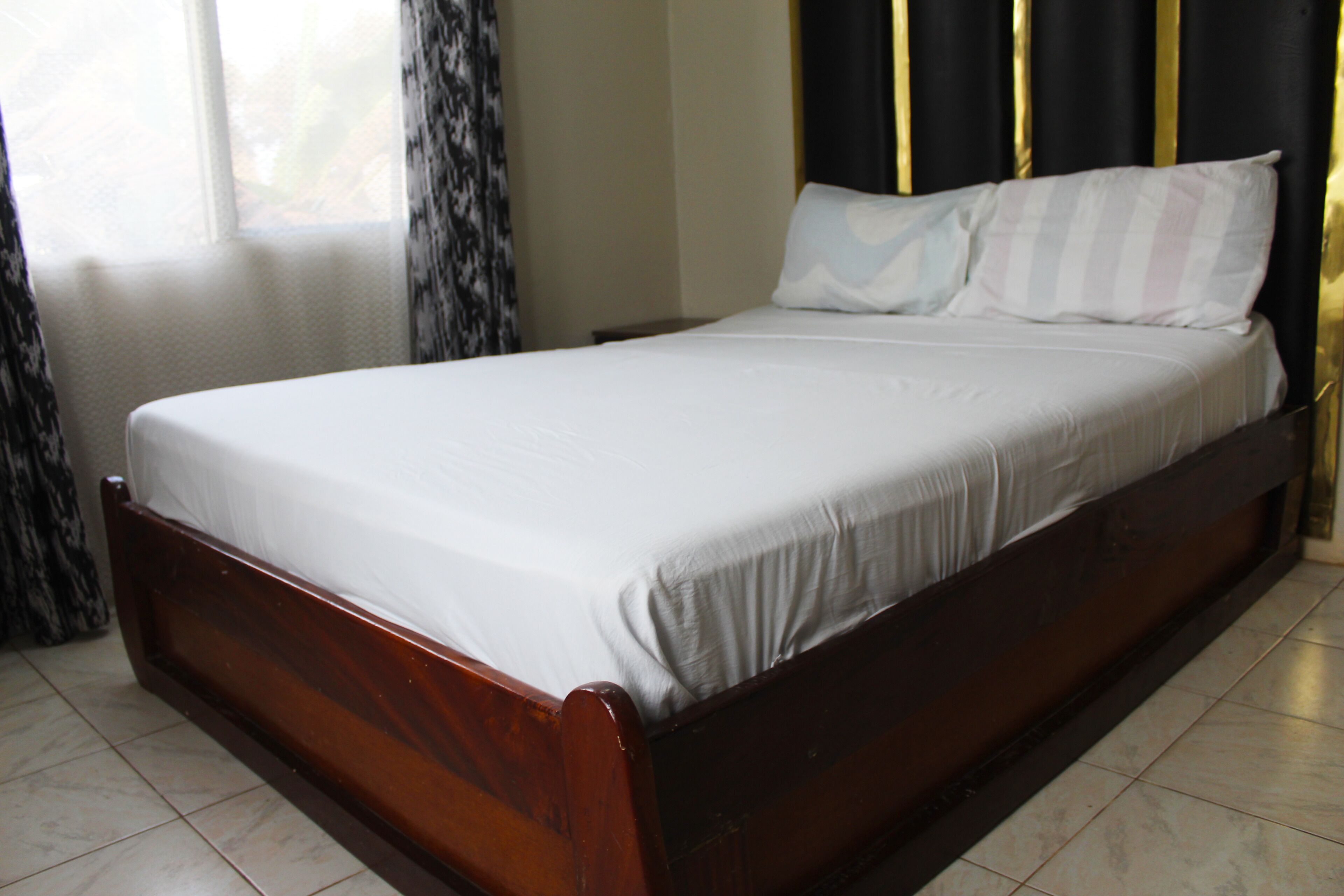 A single bed with a wooden frame and white bedding in a room with tiled flooring and a window with curtains.