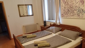 Una cama doble con ropa de cama gris y dos almohadas, una mesita de noche con lámpara, un radiador, un espejo y una ventana con cortinas.