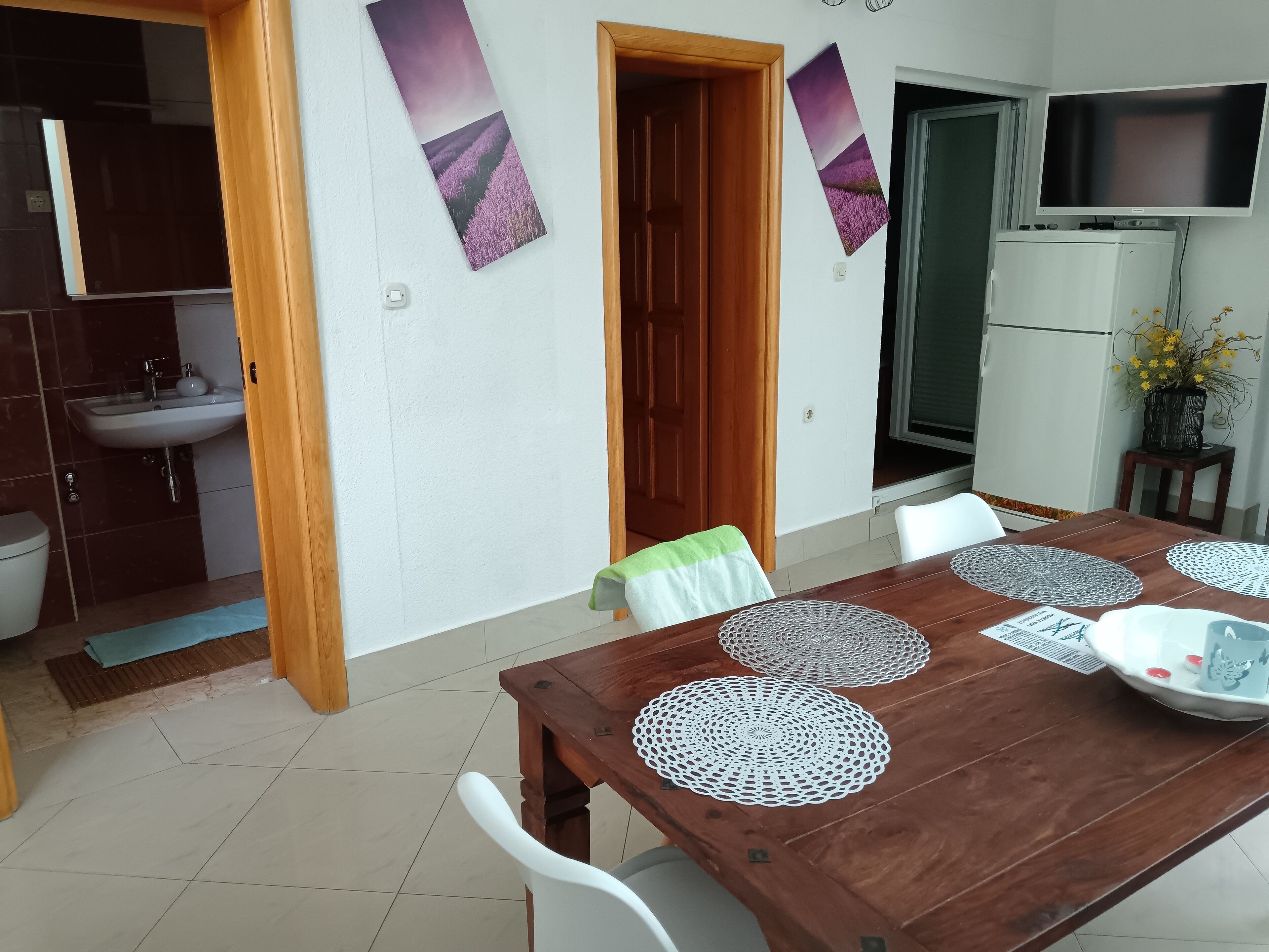 A dining area with a wooden table, white chairs, and decorative doilies. There is a kitchen with a sink and cabinets visible in the background.