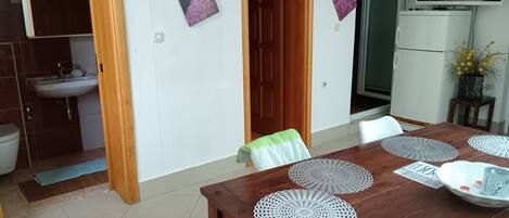 A dining area with a wooden table, white chairs, and decorative doilies. There is a kitchen with a sink and cabinets visible in the background.