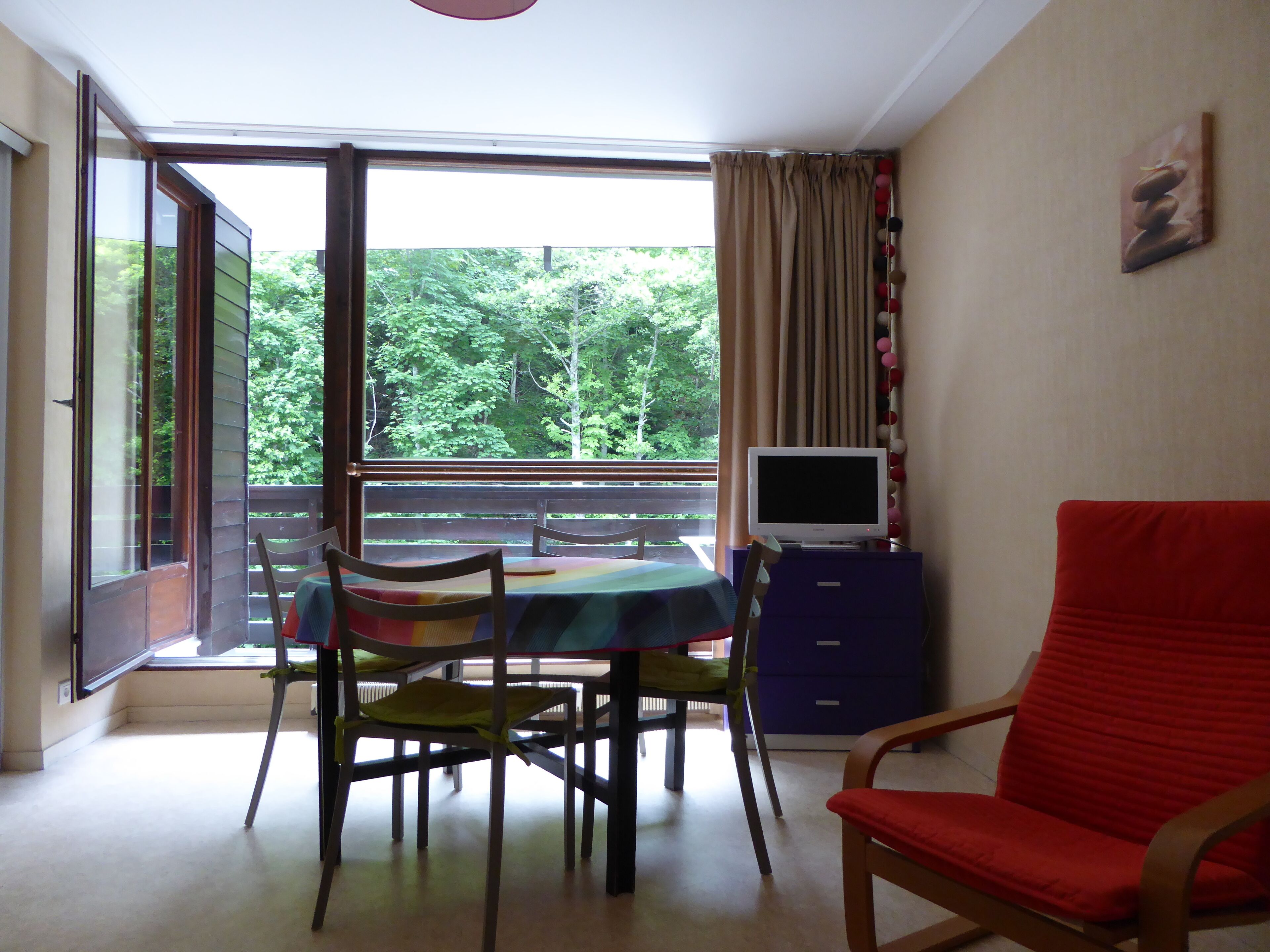 A room with a balcony, a table with chairs, a red chair, a television on a cabinet, and a painting on the wall.