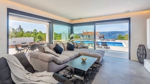 A modern living room with a sectional sofa, a coffee table, and a view of a swimming pool and mountains. - Villa QUADRA with heated pool (Pinezići)