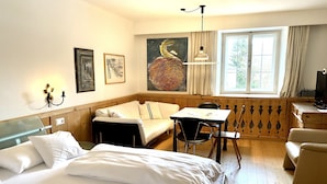 A hotel room with a bed, a sofa, a table, and a desk. There is a window with curtains, a painting on the wall, and a lamp. - Romantic studio with garden, SWIMMING POOL, mountain railways for free (Telfes)