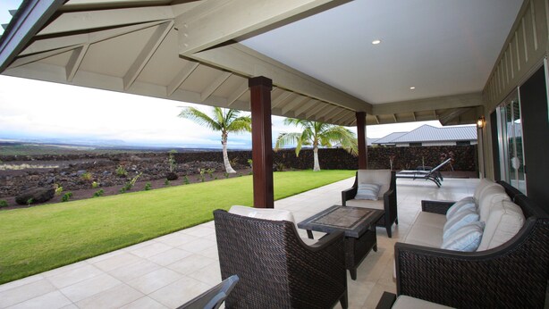 A covered patio with wicker furniture, a view of a landscaped garden, and a stone wall. - Family Friendly Home with Mountain Views, Private Lanai, and Beach Accessories (Waikoloa Village)