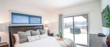 Un dormitorio con cama, mesita de noche, lámpara, ventana con vistas y puerta corredera a un balcón.