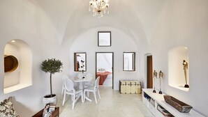 Un vaste espace de vie comprenant une table à manger, des chaises blanches, un canapé et une table basse. On y trouve également une plante en pot, un lustre et une console blanche agrémentée d’objets décoratifs.