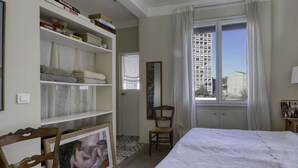 Une chambre à coucher avec un lit, une chaise en bois, une bibliothèque et une fenêtre donnant sur un immeuble.