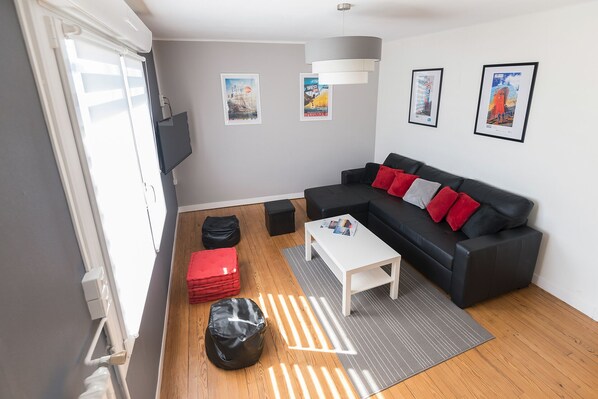 A modern living room with a black sofa, red and grey cushions, a white coffee table, and a grey rug. There are posters on the wall and a black bag on the floor.