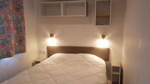 Una cama bien hecha con colcha blanca, dos mesitas de noche con lámparas, y una estantería en la pared sobre la cama.