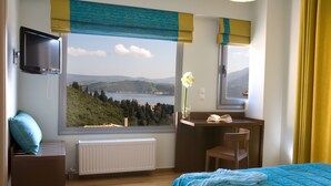 A bedroom with a large window, a bed, a desk, a chair, a radiator, and a TV.