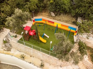 An aerial view of a playground with slides and climbing structures, surrounded by trees and a stone wall.