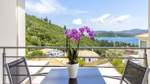 A balcony with a potted orchid, a table, and chairs, overlooking a coastal landscape with houses and greenery.