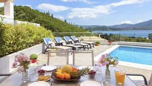 A dining table with a fruit basket, plates, cutlery, and a glass of orange juice. There are lounge chairs and a pool in the background.